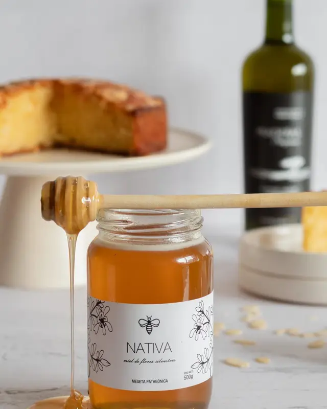 Torta cortada sobre un pie crema junto a un frasco de miel Nativa con cuchara mielera